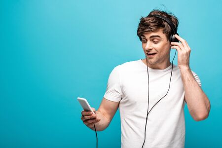 handsome man listening music in headphones and using smartphone on blue background