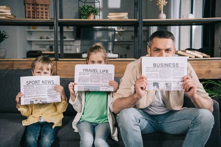 cheerful father and kids holding newspapers while sitting on sofa at homeの写真素材
