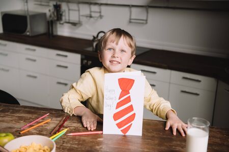 cute boy showing fathers day greeting card with drawn tie while sitting at kitchen table and smiling...