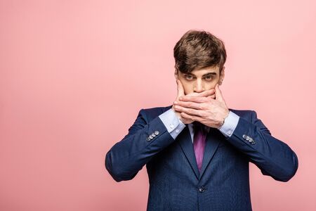 serious businessman covering mouth with hands while looking at camera on pink backgroundの写真素材