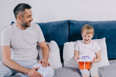 adorable boy holding gift box and fathers day greeting card while sitting on bed near happy fatherの写真素材