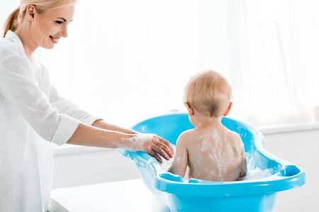 attractive and smiling mother washing toddler son in blue baby bathtubの写真素材