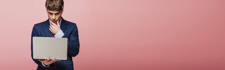 panoramic shot of thoughtful businessman using laptop on pink backgroundの写真素材