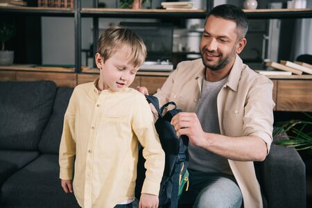 smiling dad helping cute boy putting on backpack at homeの写真素材