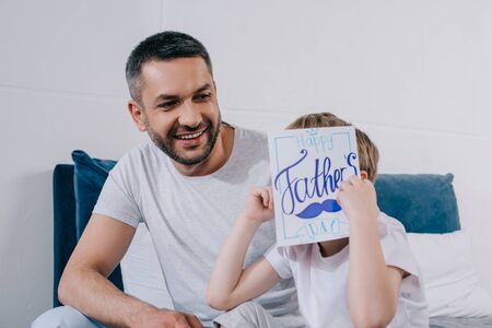 cute boy covering face with fathers day greeting card while sitting near happy fatherの写真素材