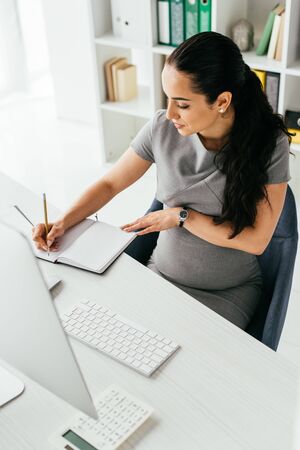 high angle view of pregnant woman sitting behind white table with computer and keyboard and making notes in notebookの写真素材