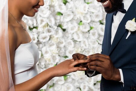 cropped view of cheerful african american man putting wedding ring on finger of bride near flowersの写真素材