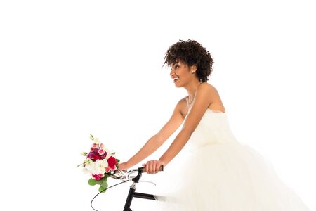 low angle view of attractive african american bride in wedding dress holding flowers while riding bicycle isolated on whiteの写真素材