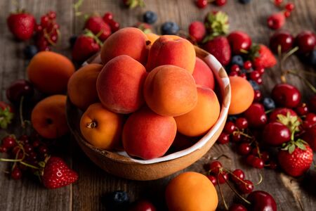 close up view of ripe delicious seasonal berries scattered around bowl with apricots on wooden tableの写真素材