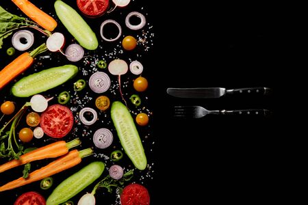 top view of fresh vegetable slices with salt near knife and fork isolated on black with copy spaceの写真素材