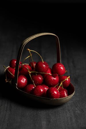 red tasty wet cherries in metal basket on wooden dark table isolated on blackの写真素材