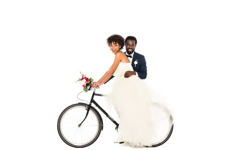 happy african american bridegroom near cheerful bride riding bicycle isolated on whiteの写真素材