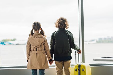 back view of kids holding hands, standing near window in waiting hall in airportの写真素材