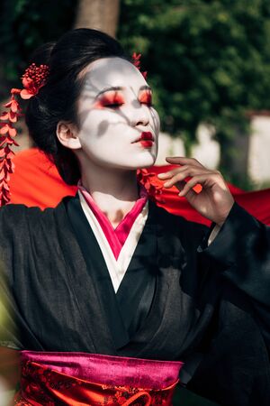 selective focus of tree branches and beautiful geisha with closed eyes and red cloth on background in sunlightの写真素材