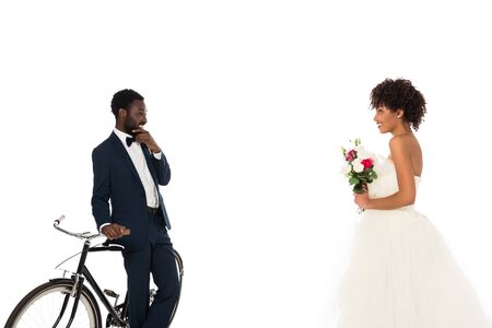 african american bridegroom standing near bicycle and looking at beautiful bride with flowers isolated on whiteの写真素材
