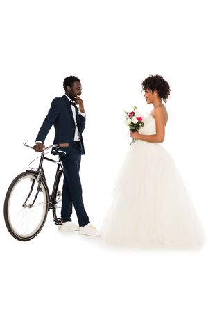african american bridegroom standing near bicycle and looking at cheerful bride with flowers isolated on whiteの写真素材