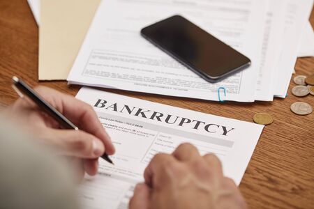 partial view of businessman filling in bankruptcy form at wooden table with smartphoneの写真素材