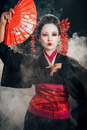 beautiful geisha in black kimono with flowers in hair holding hand fan and gesturing in smokeの写真素材