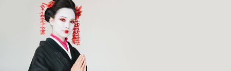 smiling beautiful geisha in black kimono with greeting hands isolated on white, panoramic shotの写真素材