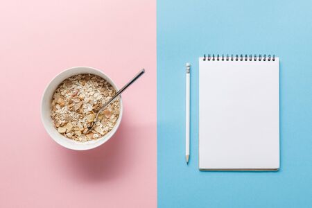 top view of breakfast cereal in bowl and blank notebook with pencil on blue and pink backgroundの写真素材