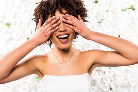 cheerful african american bride covering eyes with hands near flowersの写真素材