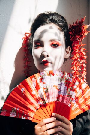 portrait of beautiful geisha with red and white makeup holding hand fan in sunlightの写真素材
