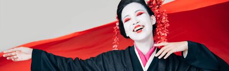 happy beautiful geisha in black kimono with red cloth on background isolated on white, panoramic shotの写真素材