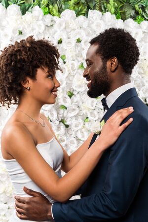 happy african american bride hugging handsome bearded bridegroom near flowersの写真素材
