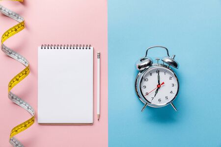top view of measuring tape and blank notebook with pencil on blue and alarm clock on pink background