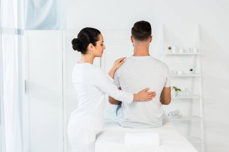 back view of man sitting on massage table near brunette healerの写真素材