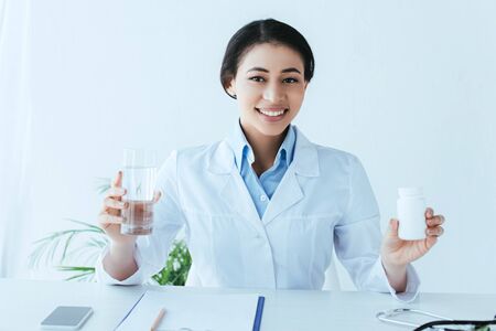 cheerful latin doctor holding pills container and glass of water while sitting at workplace and smiling at cameraの写真素材