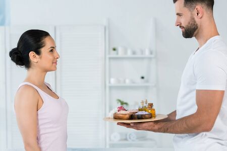 handsome masseur holding tray with sea salt and oil in bottles near attractive womanの写真素材