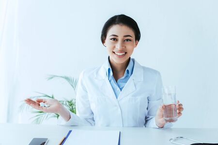 cheerful latin doctor with handful of pills and glass of water sitting at workplace and smiling at cameraの写真素材