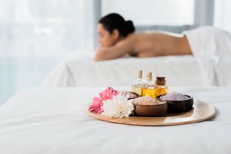 selective focus of tray with bottles of oil, bowls with sea salt and flowers near brunette woman in spa centerの写真素材