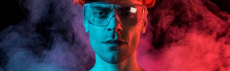 panoramic shot of serious fireman in hardhat and protective goggles in red smoke on black