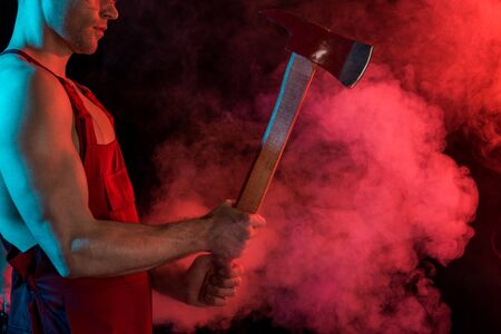 cropped view of shirtless fireman holding flat head axe in red smoke on blackの写真素材