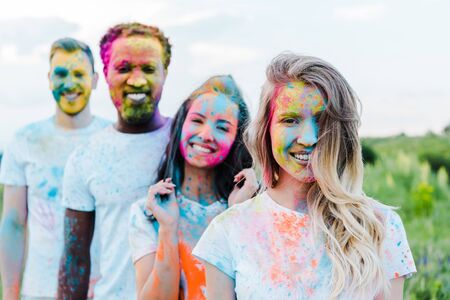 selective focus of happy woman near multicultural friends with holi paints on faces