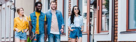 panoramic shot of happy girls with hands in pockets walking with cheerful multicultural friends