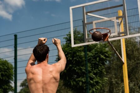 back view of shirtless basketball player throwing ball in basket at basketball courtの写真素材
