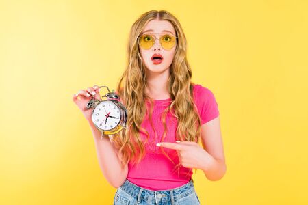 beautiful surprised girl in sunglasses pointing with finger at alarm clock On yellow