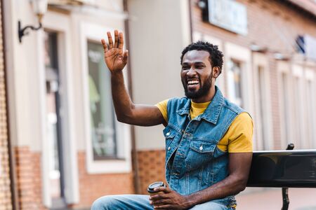 happy african american man waving hand while holding paper cup