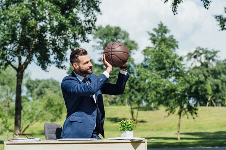 young businessman holding basketball while standing in park near table with flowerpotの写真素材