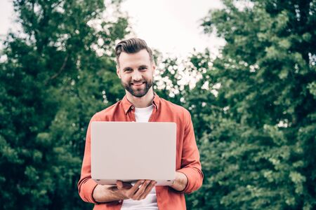 young man standing in park, holding laptop and looking at cameraの写真素材