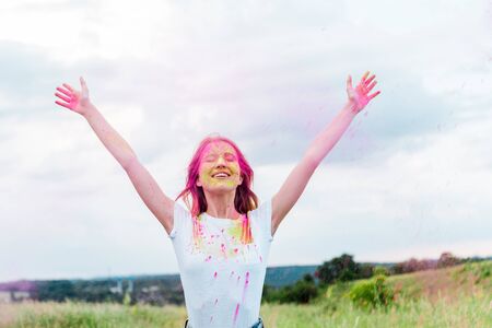 happy woman with closed eyes and pink holi paint on outstretched hands smiling outdoors