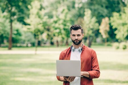 young man standing in park with laptop in hands and looking at cameraの写真素材