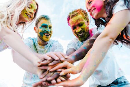 low angle view multicultural friends with holi paints on faces putting hands togetherの写真素材