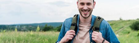 panoramic shot of happy young man standing with backpack and looking at cameraの写真素材