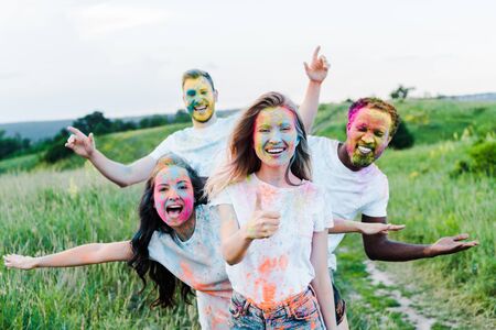 selective focus of woman showing thumb up near multicultural friends with holi paints on faces
