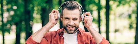panoramic shot of man putting headphones on head,  smiling and looking at camera