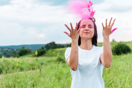 happy young woman standing with closed eyes and throwing in air pink powderの写真素材
